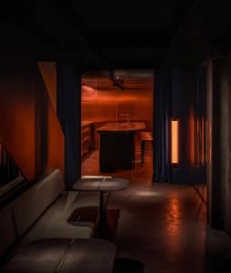 Dimly lit bar or restaurant space with a dining table, chairs, and a kitchenette, featuring wooden walls and a cozy ambiance