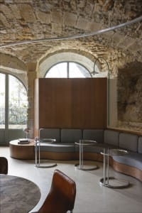 Cozy living area with stone walls, wooden furniture, and a large window, featuring a couch and several chairs around a central dining table