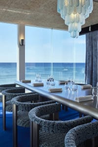 Dining area with a metal table and glass top, surrounded by chairs with cushions, overlooking the ocean