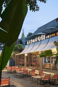 A modern Asian restaurant patio with tables, chairs, umbrellas, and potted plants, featuring a warm atmosphere with orange and white colors and natural and artificial lighting