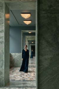 A woman walks down a luxurious hallway with a marble floor, lit by overhead lights and reflected in a nearby mirror