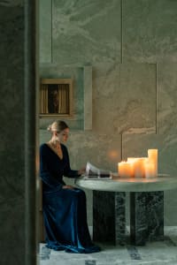A woman sits at a table in a dimly lit room with a stone wall, surrounded by candles and a vase, with two chairs and a book