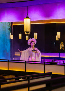 A woman in traditional attire stands on a stage in a purple-themed restaurant, surrounded by bowls and bottles, holding a plate of food and wearing a flower-adorned hat