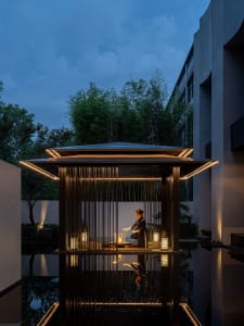 A serene outdoor setting with a small wooden structure resembling a pagoda or traditional Japanese house, featuring a woman meditating in the peaceful environment