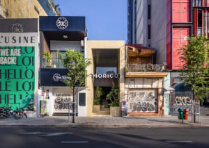 A lively urban street scene with a building facade, potted plants, and two benches in front, creating a welcoming atmosphere