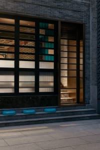 A modern building interior with a large window and glass door, featuring three blue stools outside the entrance, allowing natural light to enter the space