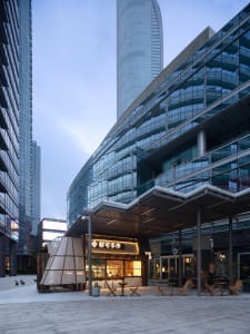Lively urban street scene with a glass building facade, outdoor seating area, and people enjoying the atmosphere