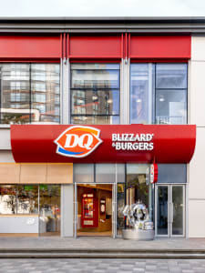 Exterior view of a modern restaurant with a red roof, multiple windows, and a large sign reading DQ Blizzard Burgers, with two clocks on the side
