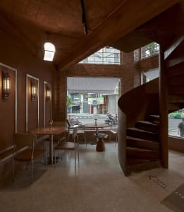 A modern cafe with a spiral staircase, brown and red tones, and cozy lighting from table lamps