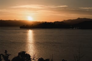 A serene lake scene at sunset with mountains in the background, warm hues of orange and pink in the sky, and two boats on the water
