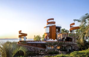 A multi-story house near water with a mix of stone, wood, and glass materials, showcasing modern elements and a luxurious atmosphere