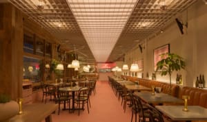 A modern restaurant interior with a long dining table, pink carpet, and potted plants, featuring wine glasses and a well-lit atmosphere
