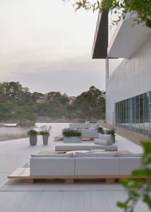 A relaxing outdoor living area with a white couch, wooden deck, and potted plants near a body of water