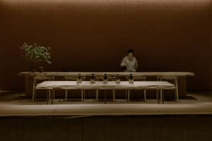 A woman stands in front of a wooden dining table surrounded by chairs and vases, in a restaurant-like atmosphere