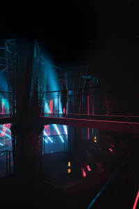 A modern interior space with a red glow, featuring a woman standing on a bridge overlooking a room with scattered chairs