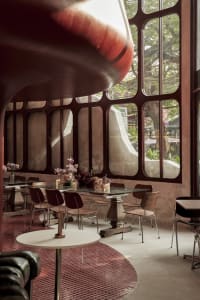 Modern restaurant interior with red walls, large windows, and natural light, featuring tables, chairs, and potted plants