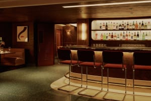 A cozy bar area with a long wooden counter and multiple stools, surrounded by comfortable seating and a well-stocked bar with bottles and a welcoming atmosphere