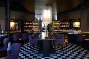 A modern bar area with a long dining table, black and white checkered flooring, and several bottles on the table