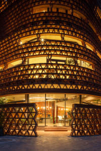 A modern building with a large wooden structure in front, featuring a seating area with chairs and potted plants both inside and outside