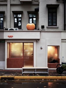 A rustic building with a brown wooden door and two benches in front, surrounded by a natural landscape with a large orange apple on the roof
