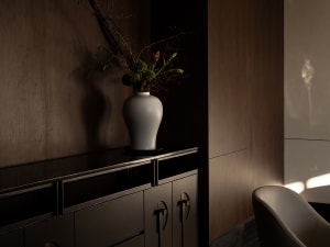 Dimly lit room with a white vase on a table, containing flowers, and a chair against the wall, with two drawers nearby