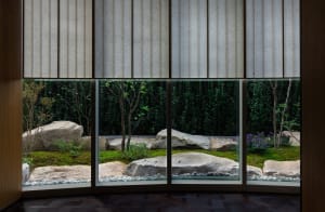 A tranquil indoor space with a large window offering a view of a garden with a pond, rocks, and plants, surrounded by trees, and natural light pouring in