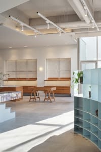 Open living area with white walls, wooden floors, and natural light from a large window, featuring chairs and potted plants
