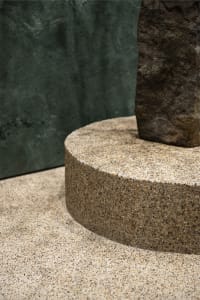A stone building with a modern design featuring a large stone column in front of a green wall, with two other columns nearby, showcasing a combination of gray and green tones