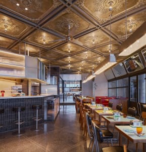 A modern commercial kitchen with a dining area featuring a table and chairs, appliances, and scattered bottles, set in a restaurant or commercial kitchen setting