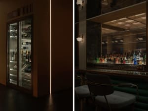 A modern bar area with a green wall, refrigerator, chairs, and bottles, with a couch and dining table in the background