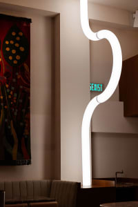 A modern room with a large white wall featuring a green sign reading sexist, a curved light, and a painting on the wall