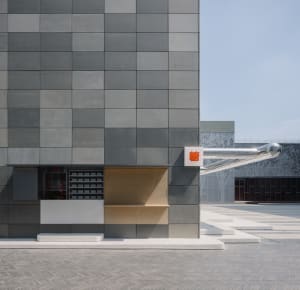 Exterior view of a modern building with a large gray wall featuring unique design elements and two signs, set against a clear blue sky