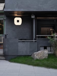 Modern building exterior with a gray wall and a large rock in front, featuring two windows and a concrete facade