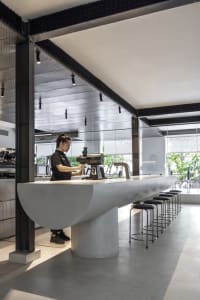 A modern cafe with a long counter and stools, surrounded by chairs and natural light from a large window, with a woman standing behind the counter