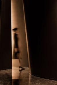 A woman walks down a wooden hallway with pillars, surrounded by two other people in a historical setting