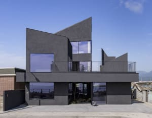 A modern house with a black exterior, large windows, and a patio area with two chairs, set against a background of water
