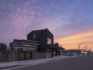A modern house with a glass wall at dusk, set in a serene residential area near a beach with a pier in the background