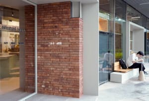 A brick wall with a textured, rustic appearance in front of a window, with two benches and a person standing nearby