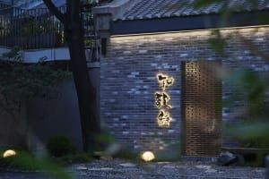 Asian-style building with a brick wall and stone facade, featuring a doorway with Chinese characters, surrounded by two trees in a calm and serene atmosphere