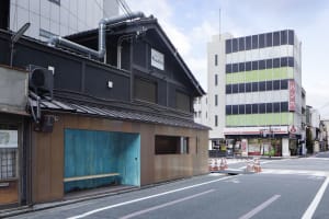 A modern building with a wooden facade and a blue roof, featuring a small shop or cafe in front, outdoor benches, and construction barriers