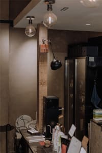 A cozy kitchen and living area with two refrigerators, a microwave, wooden beam, and wooden table, with scattered bottles and a warm atmosphere
