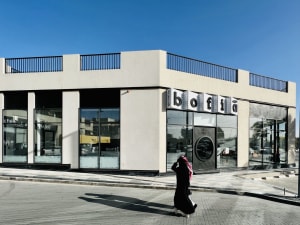 A casual street scene with a woman walking in front of a building, surrounded by parked cars, outdoor furniture, and a dining table, set against a relaxed urban backdrop