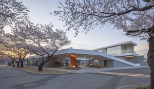 Modern building with a large glass wall, surrounded by trees and flowers, featuring a curved roof and a serene atmosphere