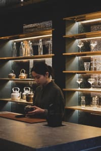 A woman stands in front of a wooden counter filled with wine glasses, cups, and bottles, surrounded by a social gathering atmosphere
