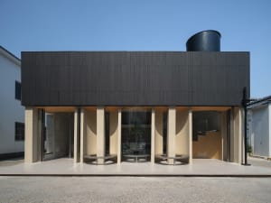 Modern building exterior with a black facade, featuring two wooden benches in front, set against a neutral background