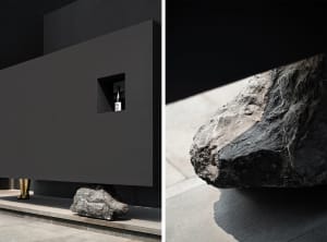 Dark modern house with a floor plan showing a kitchen area with a sink and dining table, and an architectural scene with large concrete rocks in front of a black wall