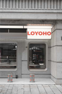 Exterior view of a modern industrial building with gray concrete walls, two large windows, and two umbrellas in front, with a person standing near one window