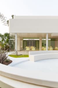 Open modern living area with white walls, floors, and chairs, and two dining tables in a bright and airy space