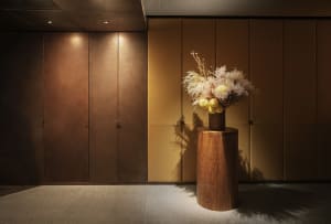 A cozy hallway with a yellow wall, a wooden vase with flowers, and a wooden stand, with an open door revealing a room beyond