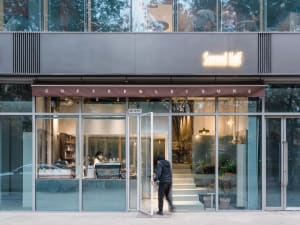 A spacious coffee shop with a glass door entrance, couches, chairs, potted plants, and scattered books, creating a cozy atmosphere for reading and relaxation
